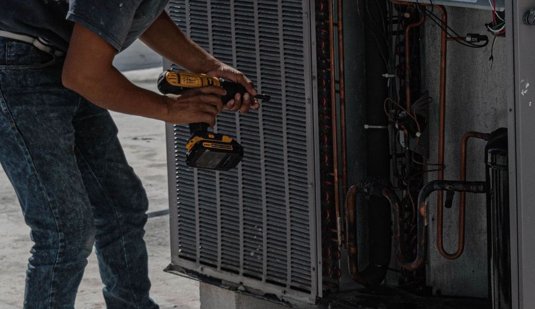 A worker in a bucket hat repairs an outdoor air conditioning unit on a rooftop.