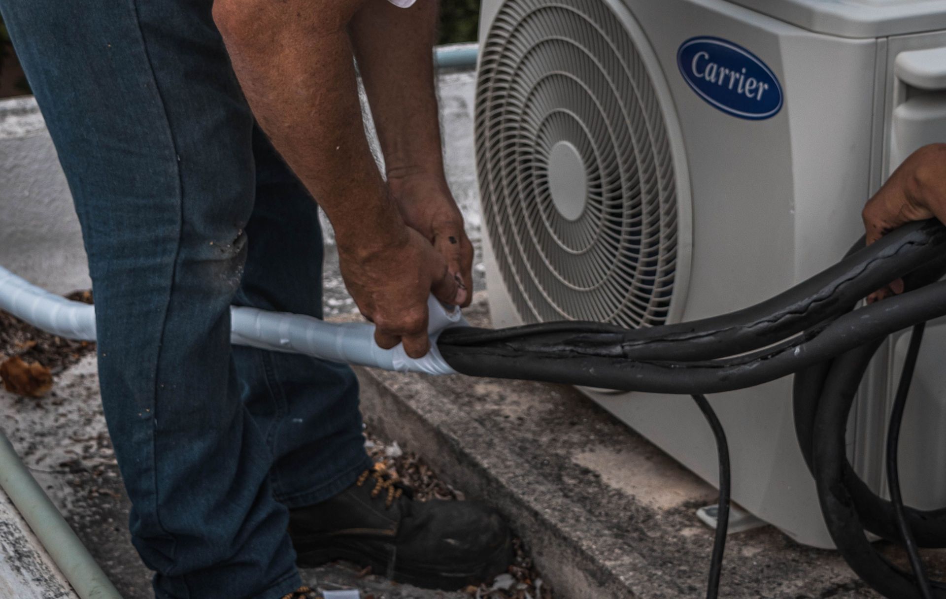 A technician performs maintenance on an outdoor air conditioning unit, focusing on hose connections.